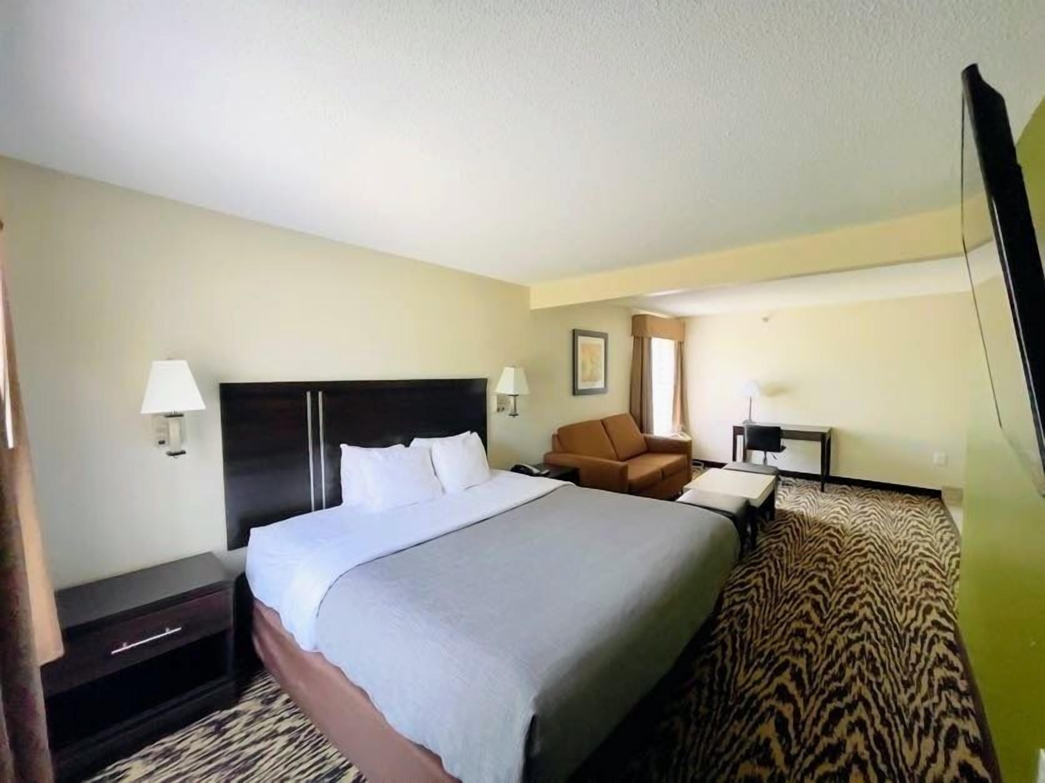 A hotel room with a large bed, a brown armchair, a desk with a chair, and a small table.