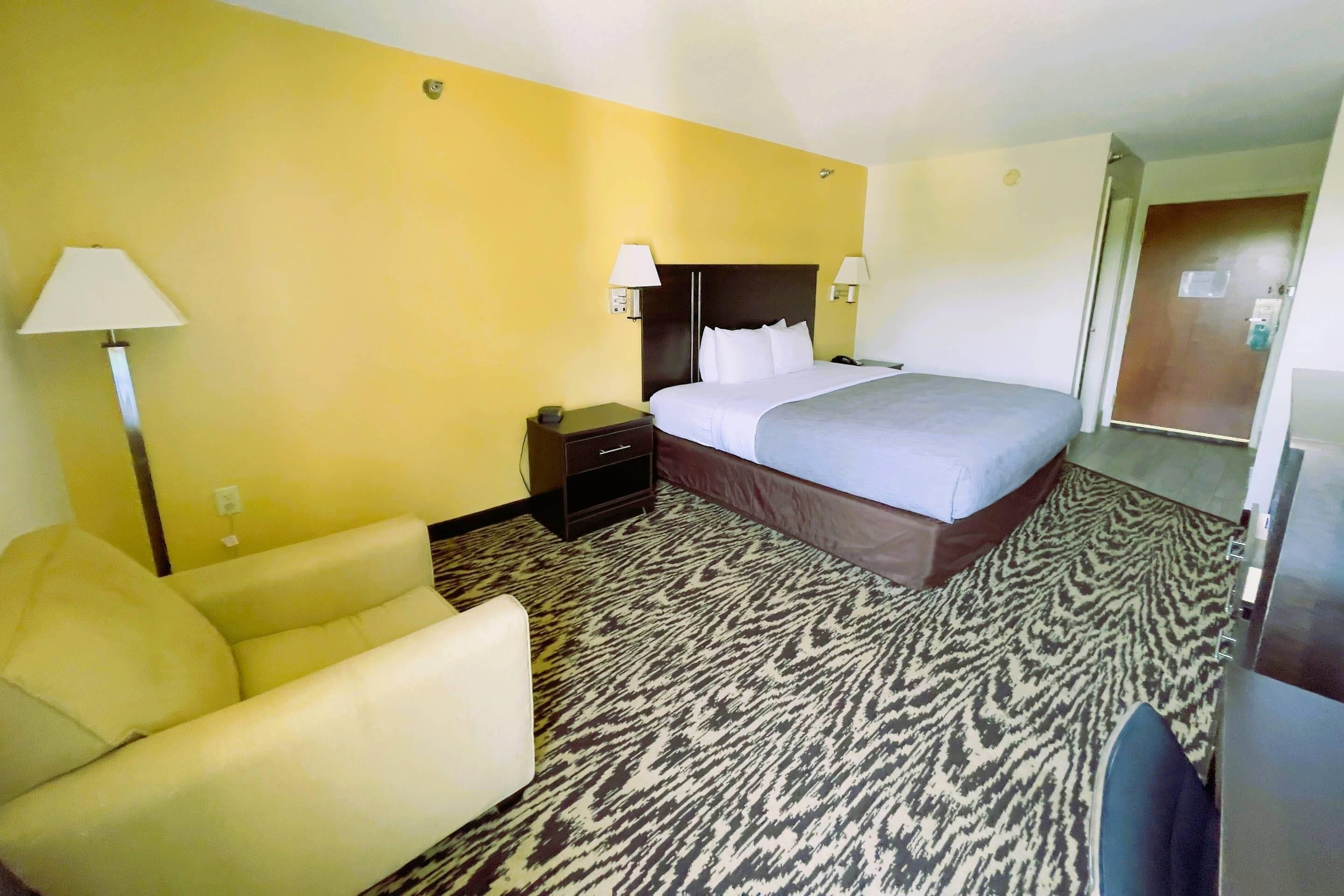 Ein Hotelzimmer mit Bett, Sessel, Nachttisch, Lampe und einem gemusterten Teppich.