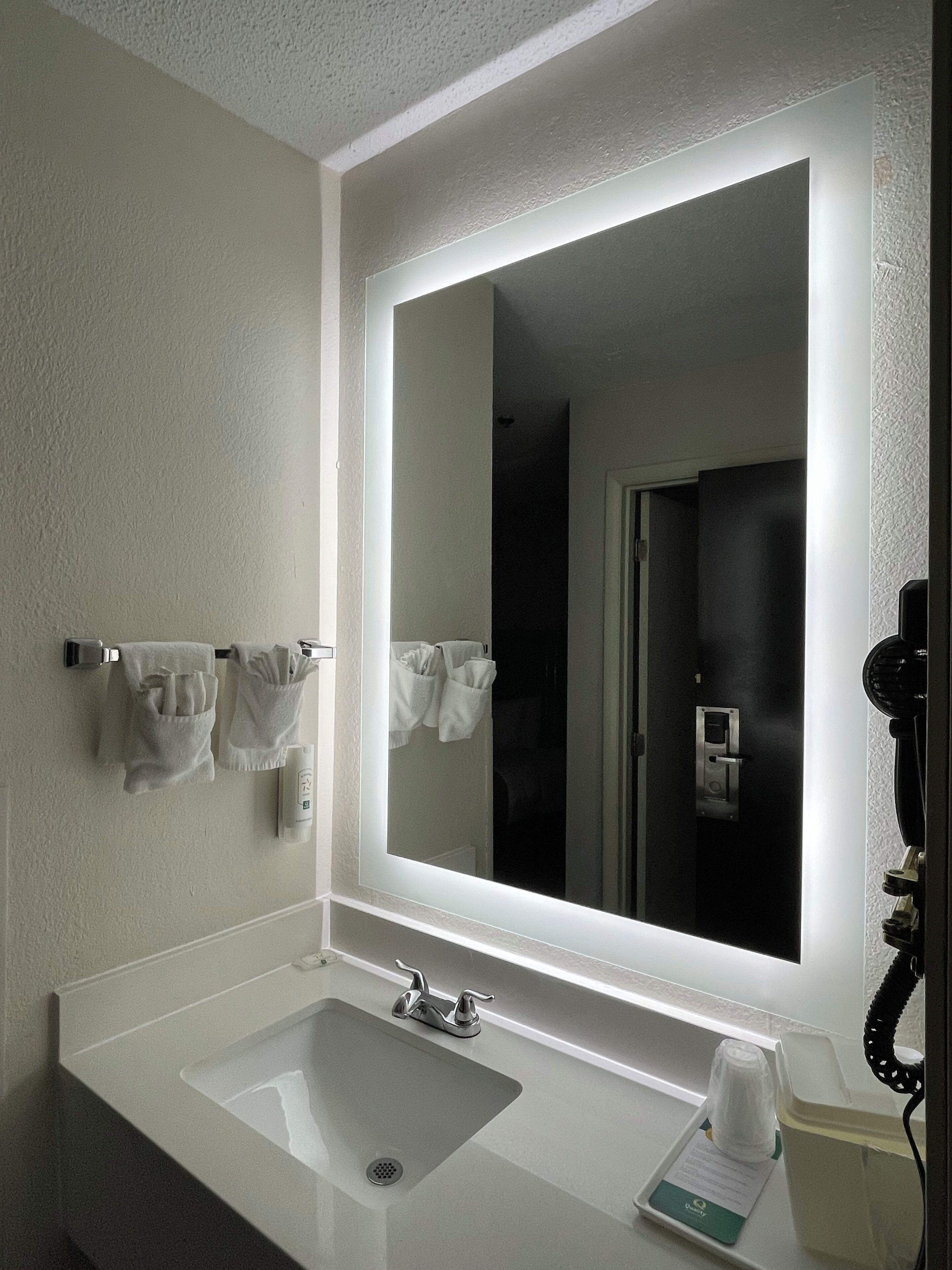Un baño de hotel con un espejo grande, un lavamanos y un perchero para toallas.