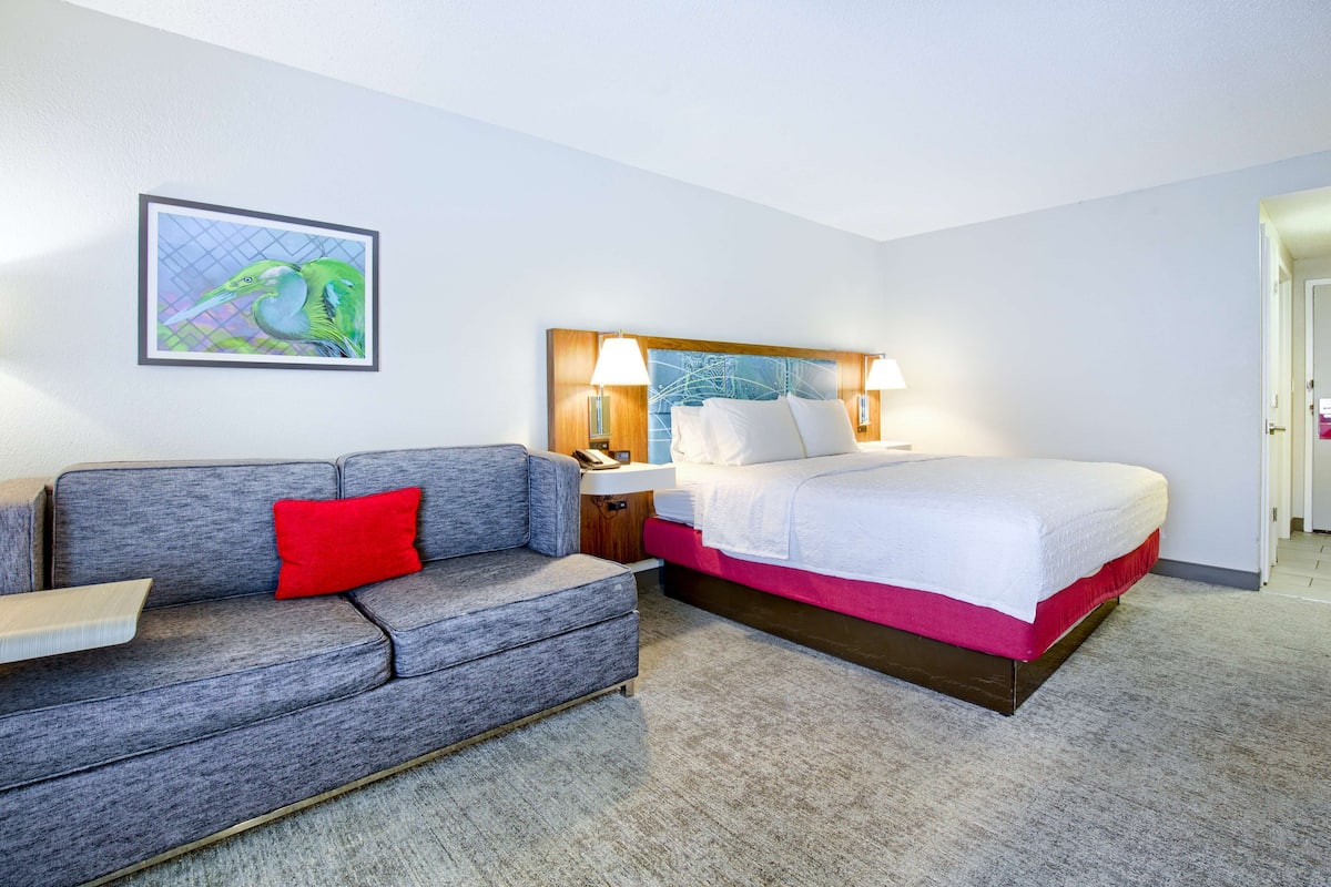 A hotel room with a bed, a gray sofa with a red pillow, a nightstand, and a wall-mounted lamp.