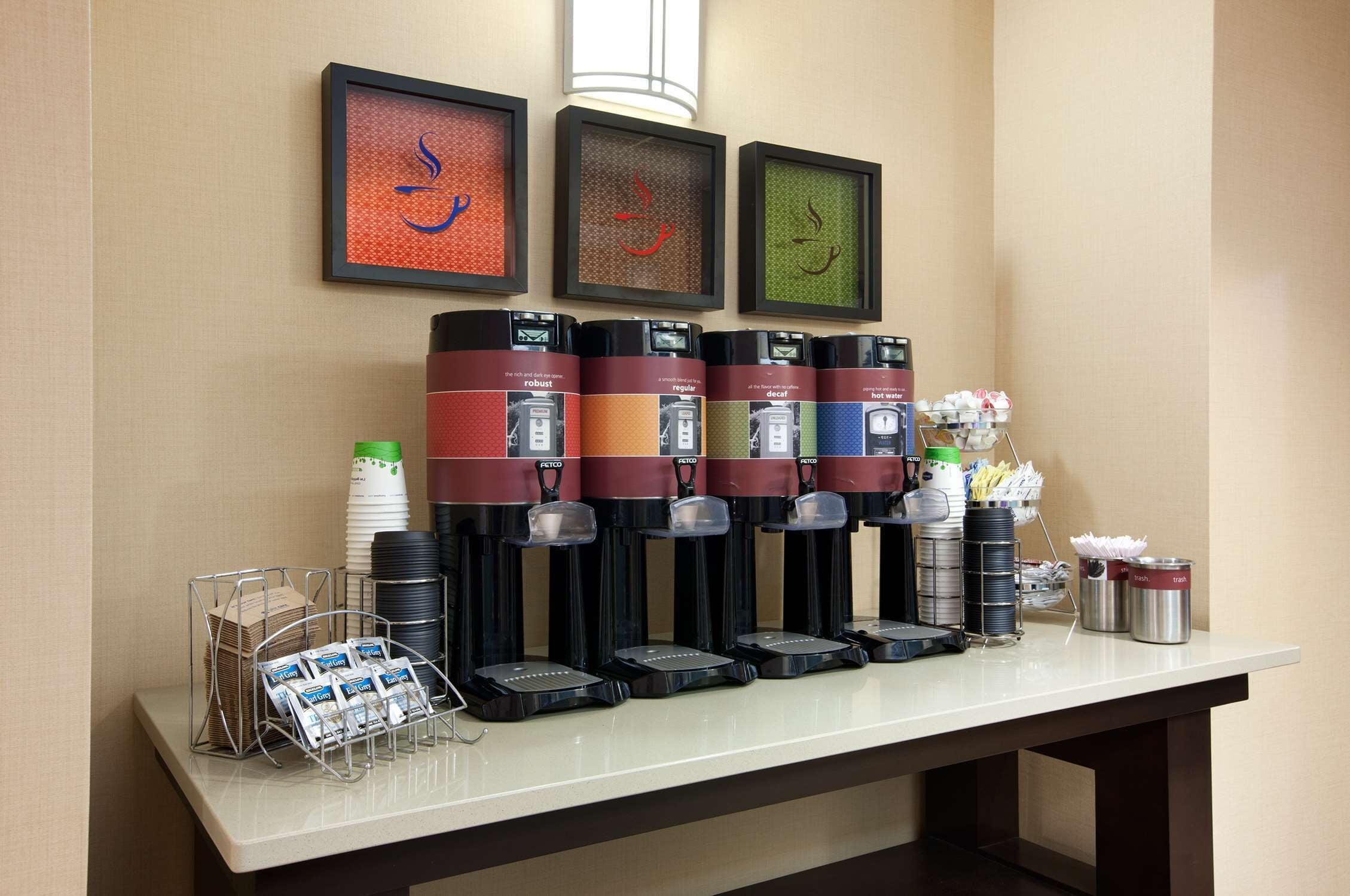 A beverage station with multiple dispensers, a stack of disposable cups, and a trash can.