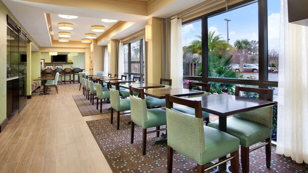 A hotel dining area with multiple tables and chairs, large windows with a view of the outdoors, and a kitchen area in the background. - Hampton Inn Slidell (Slidell)