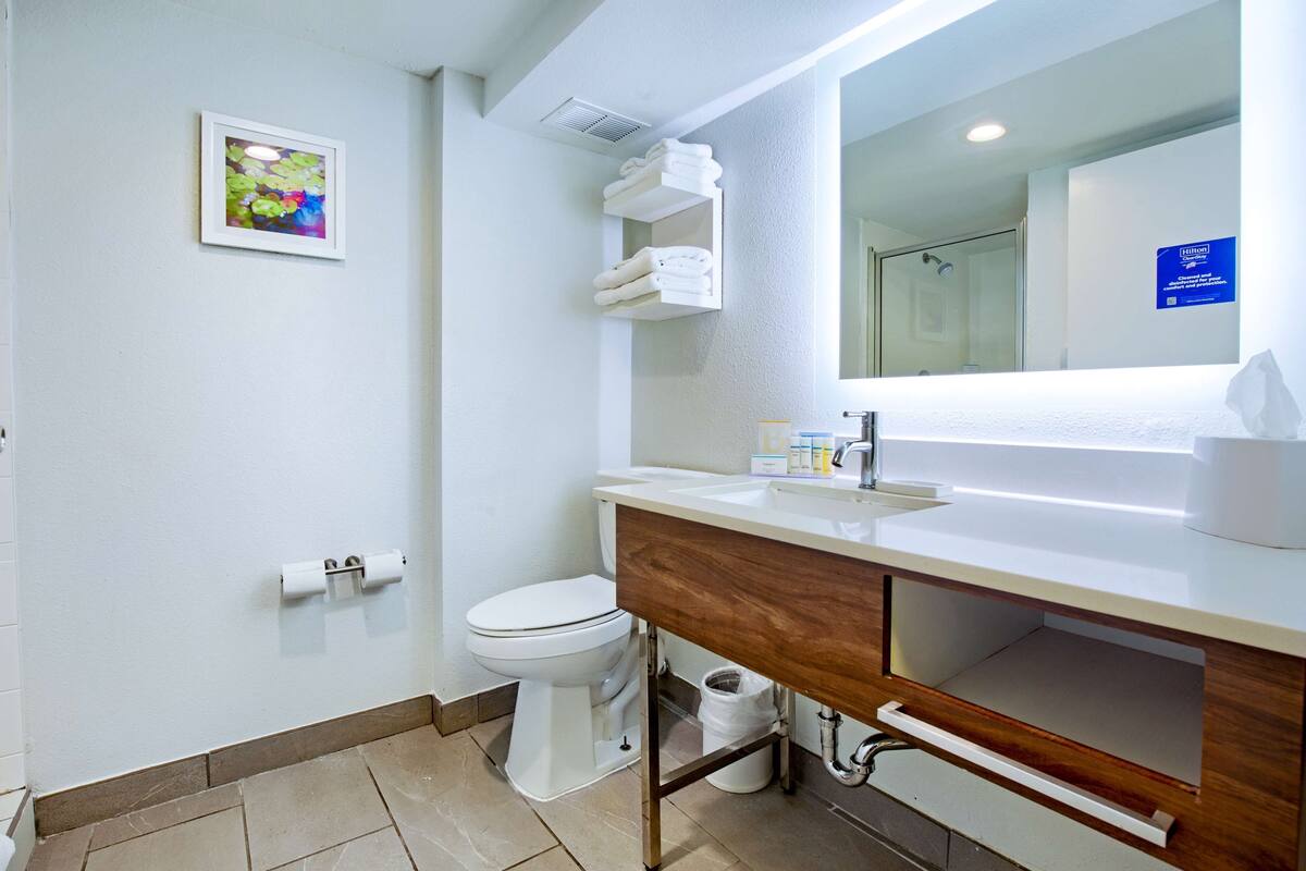 A modern bathroom with a toilet, sink, and mirror. There is a towel rack above the sink and a trash bin below.