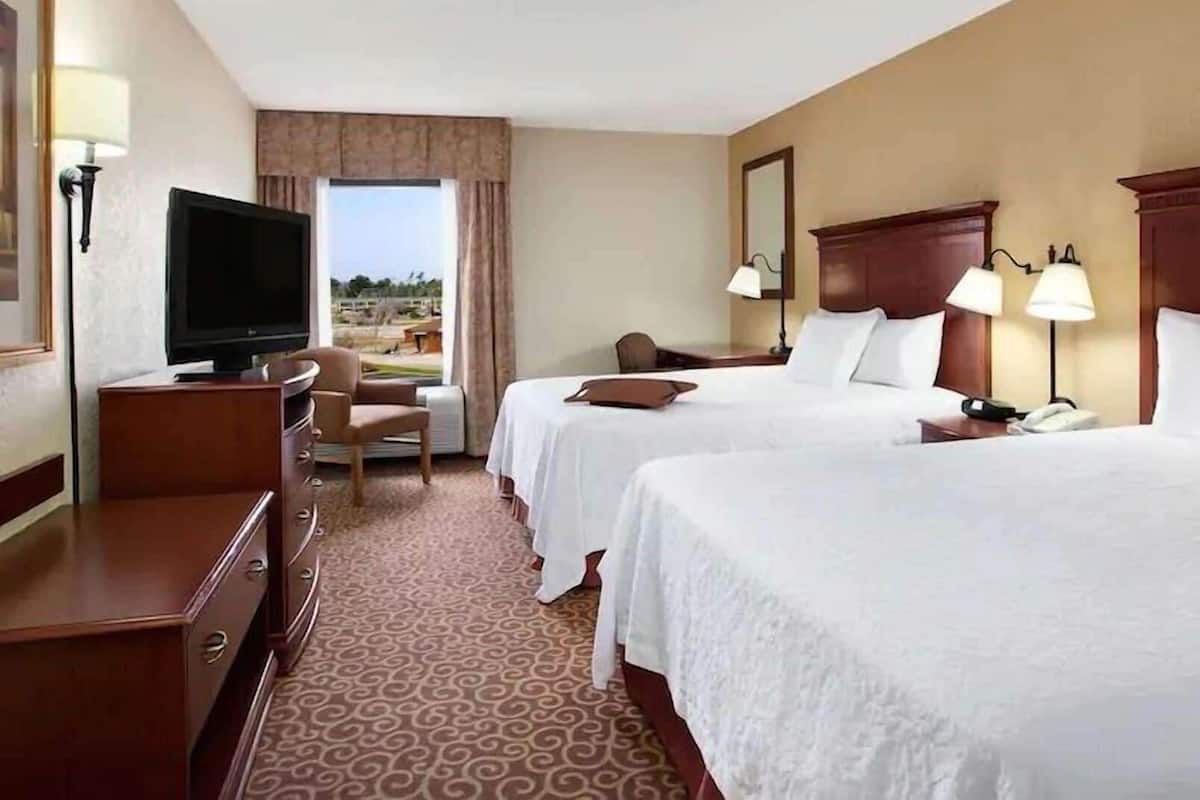 A hotel room with two beds, a TV, a desk, and a view of the outdoors.