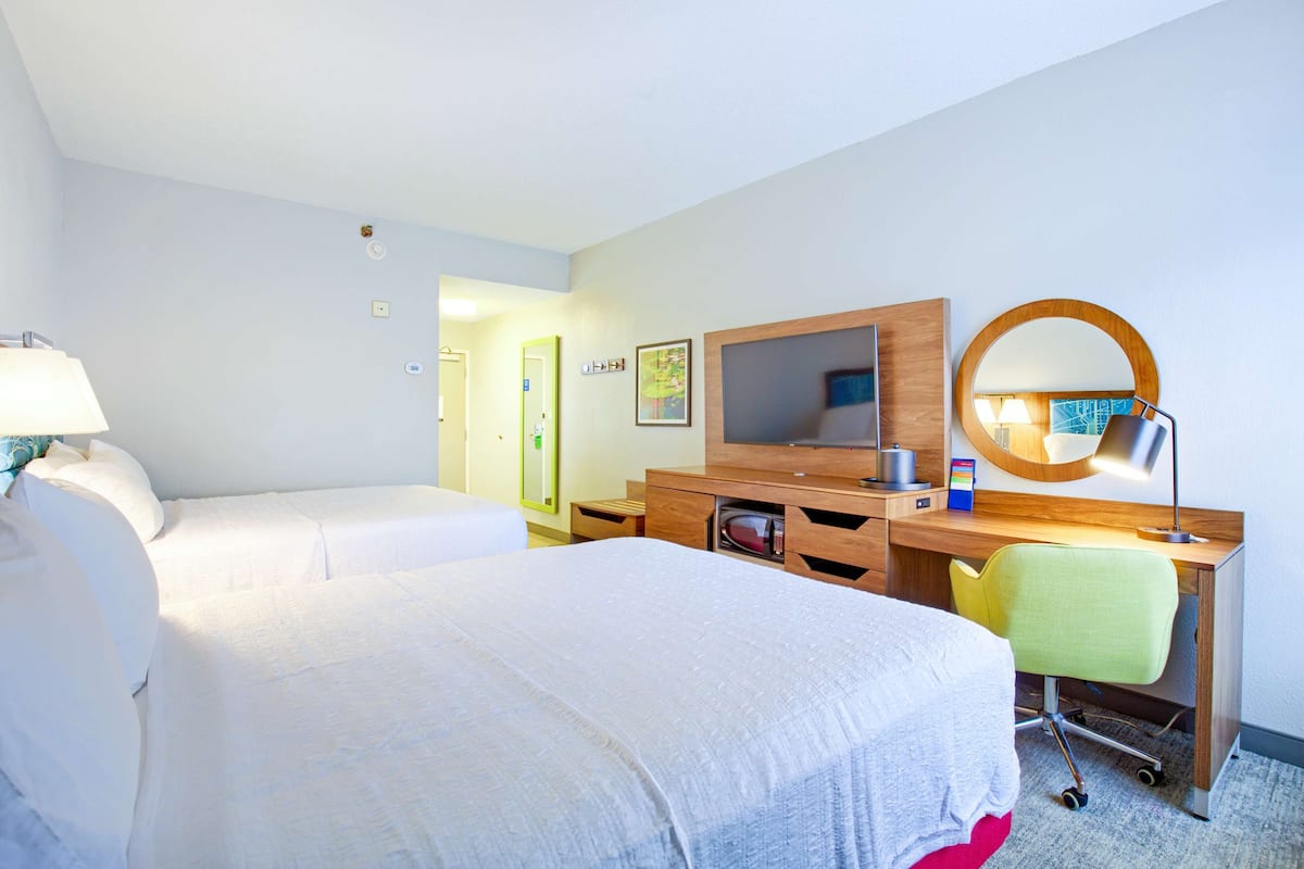 A hotel room with two beds, a desk with a mirror, a TV, and a desk lamp.