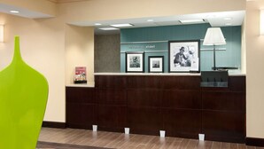A reception area with a dark wood reception desk, a green chair, and framed pictures on the wall. - Hampton Inn Slidell (Slidell)