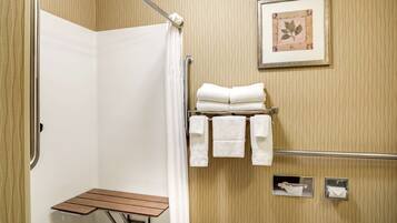 A bathroom with a shower, a wooden bench, a towel rack with towels, a framed picture on the wall, and a toilet paper holder.
