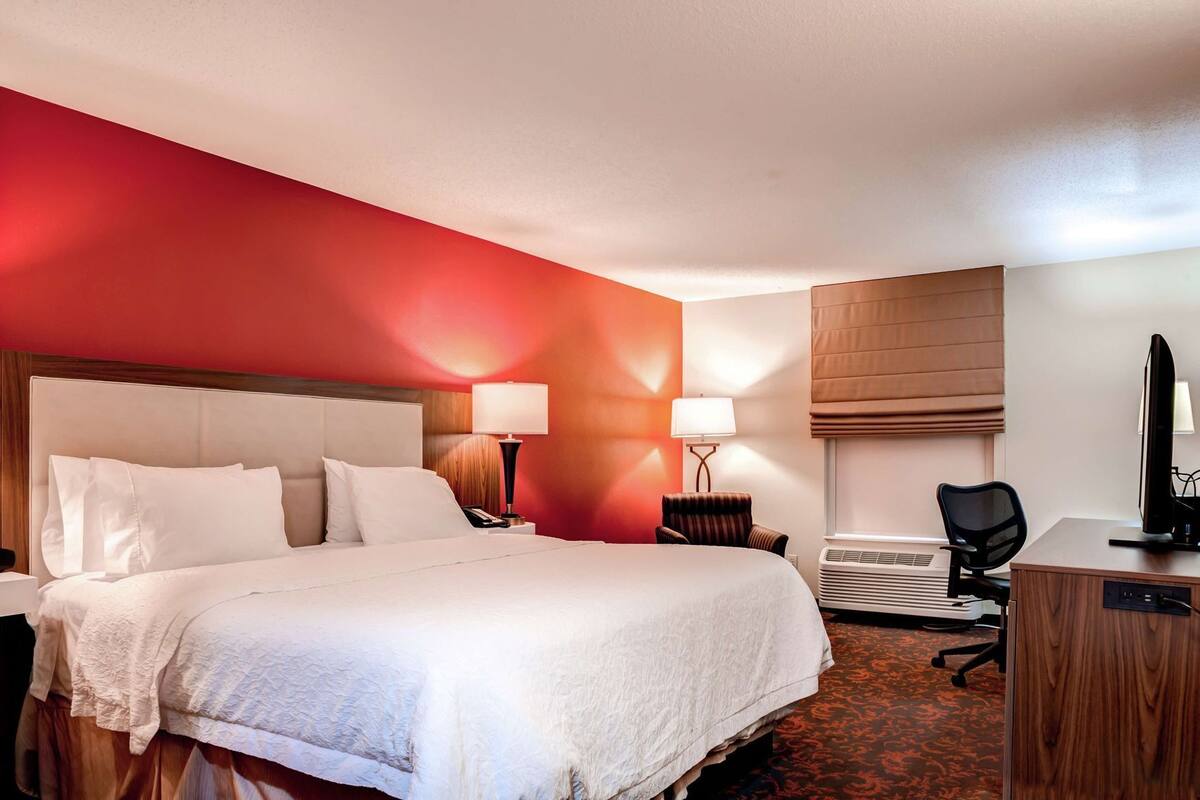 A hotel room with a large bed, a desk with a computer, a chair, a lamp, and a window with blinds.