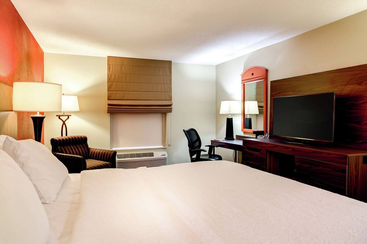 A hotel room with a large bed, two chairs, a desk with a TV, and a mirror.