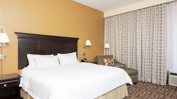 Ein Hotelzimmer mit einem großen Bett, einem Nachttisch, einem Sessel und einem Fenster mit gemusterten Vorhängen.