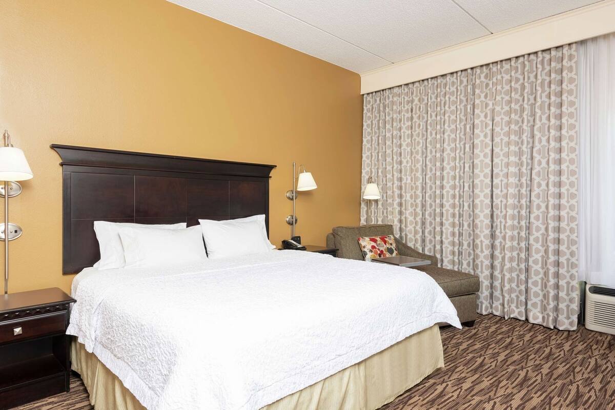 A hotel room with a large bed, a nightstand, a chair, and a window with curtains.