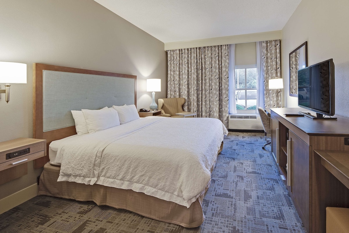 A hotel room with a large bed, a desk with a television, a chair, and a desk lamp.