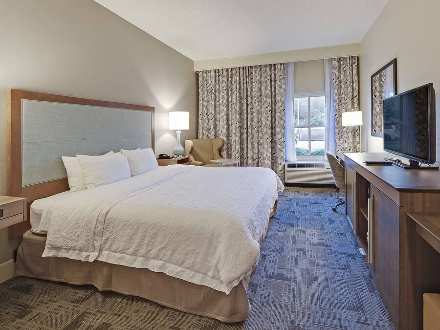 A hotel room with a large bed, a desk with a television, a chair, and a desk lamp.