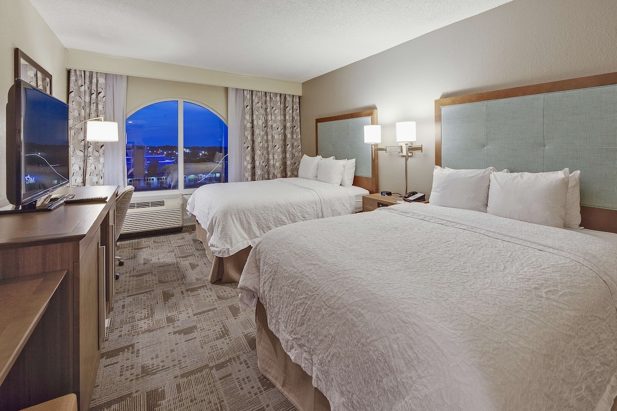 A hotel room with two beds, a desk with a TV, and a view of the city at night.