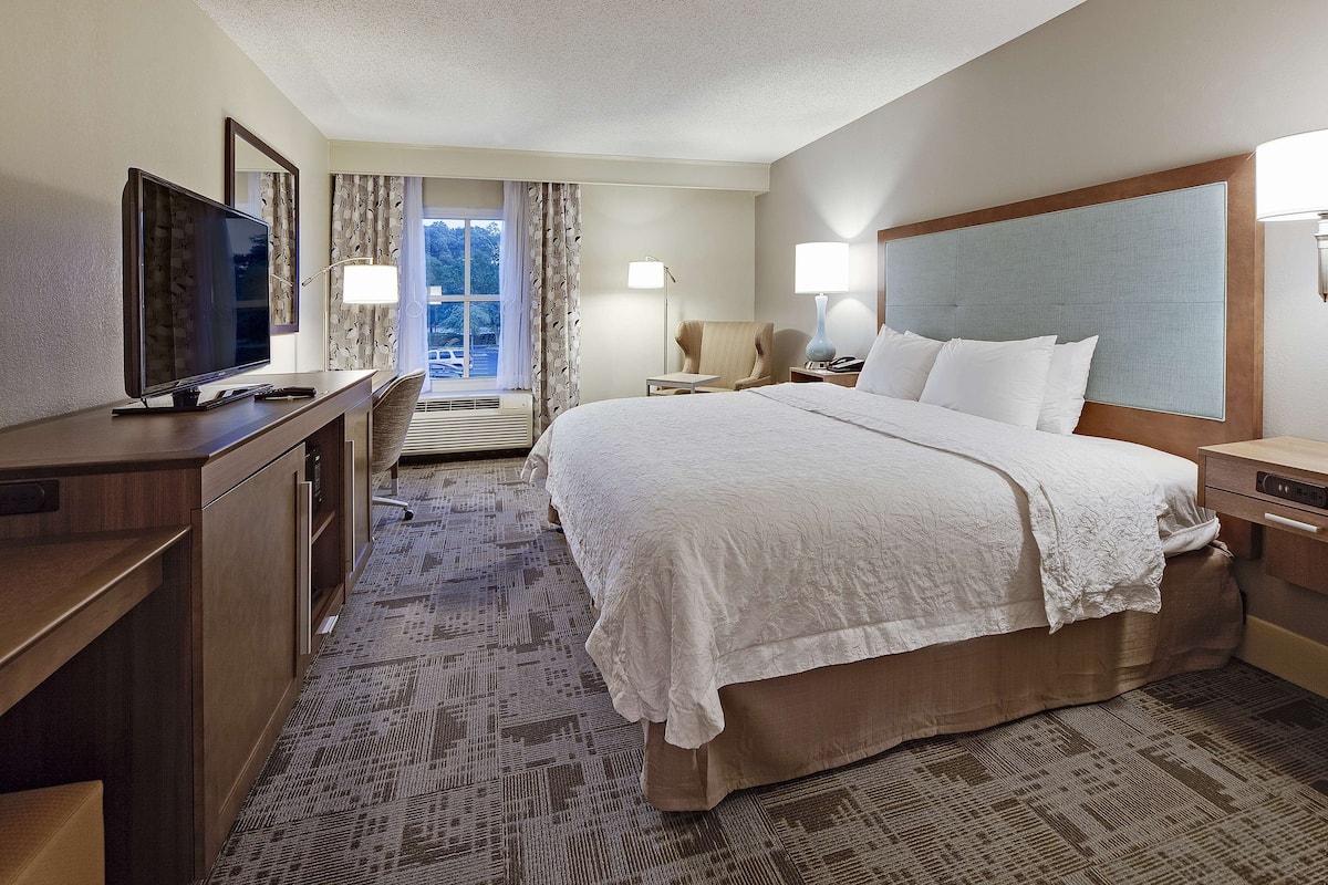 A hotel room with a large bed, a desk with a television, a chair, and a window with curtains.