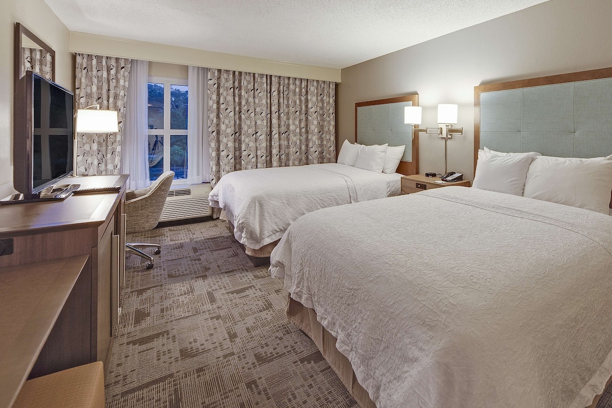 A hotel room with two beds, a desk with a TV, a chair, and a window with curtains.