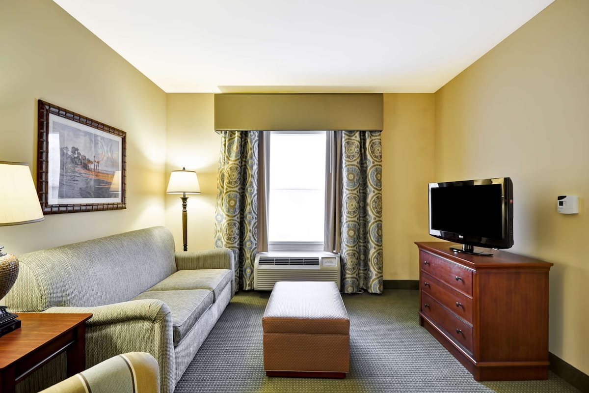 A hotel room with a sofa, a television on a wooden cabinet, a lamp, an air conditioning unit, and a window with patterned curtains.