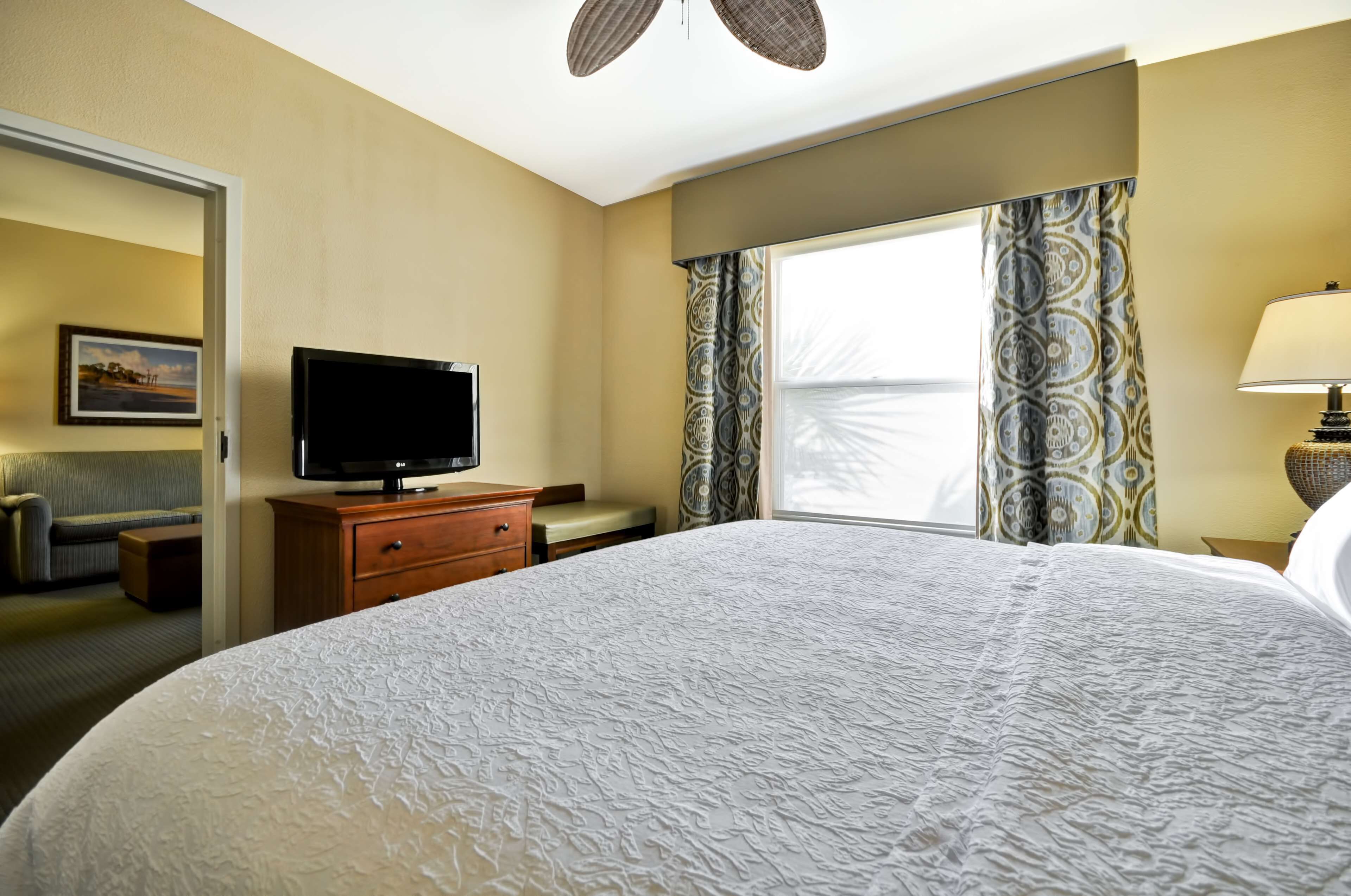 Une chambre d’hôtel avec un grand lit, un téléviseur posé sur une commode en bois, un bureau et une fenêtre ornée de rideaux à motifs.
