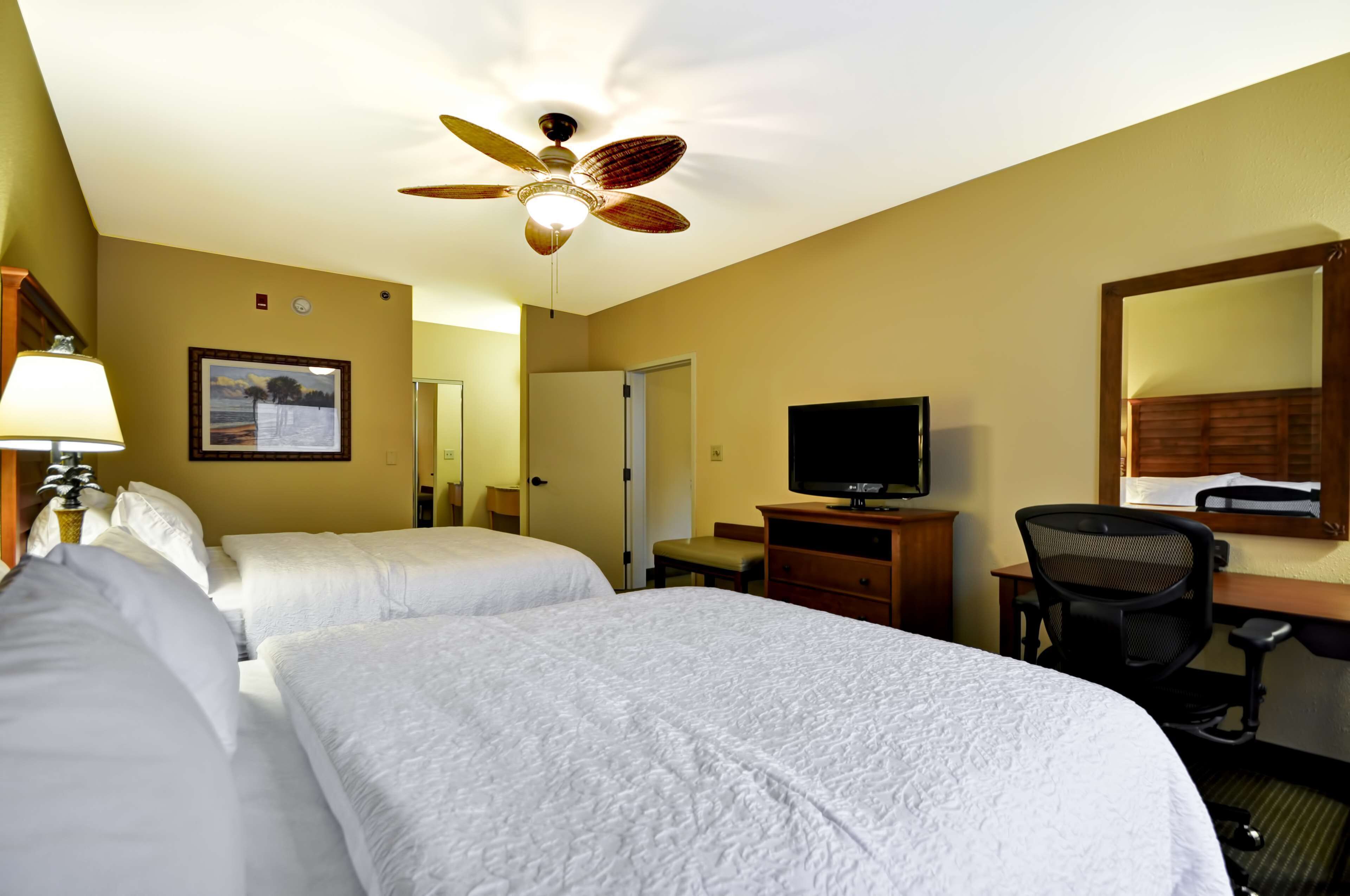 Habitación de hotel con dos camas, un escritorio con una silla, un televisor, un ventilador de techo y un espejo.