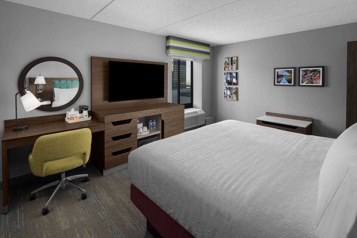 A hotel room with a bed, a desk with a chair, a TV, a mirror, a window, and a wall with framed pictures.