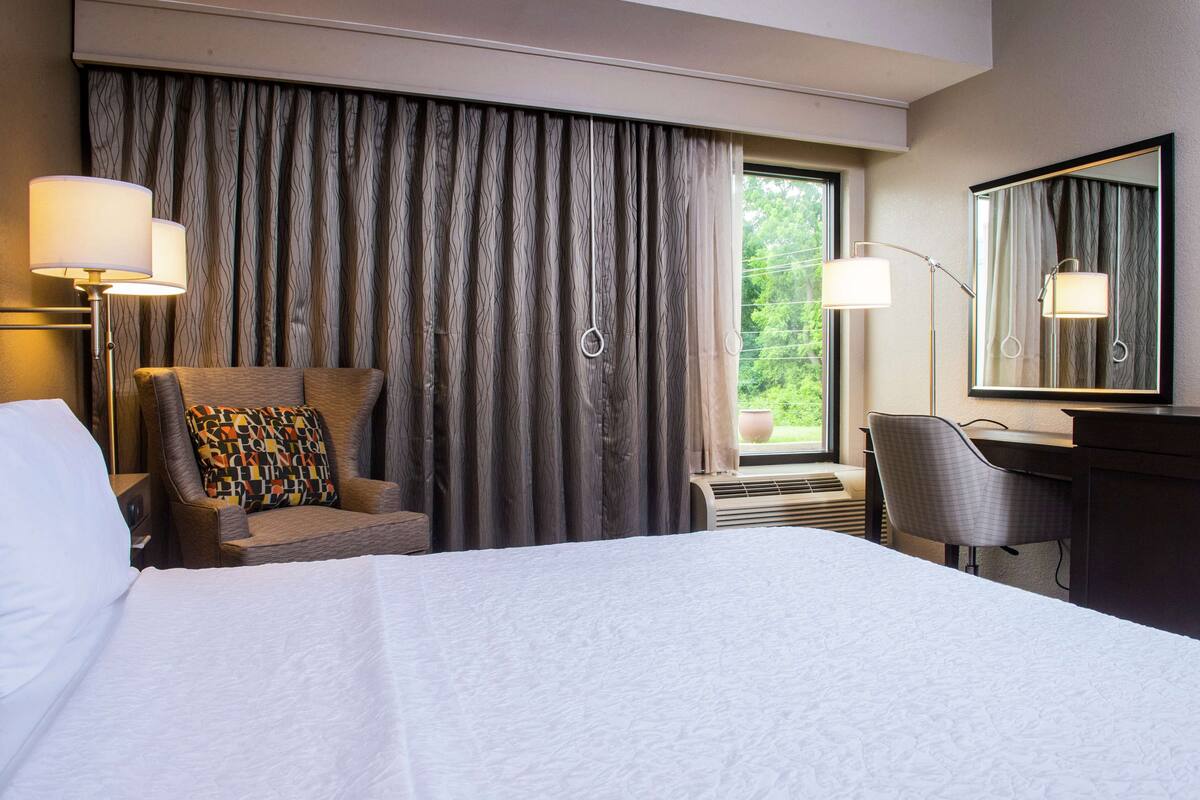 A hotel room with a large bed, a desk, a chair, a lamp, and a window with curtains.