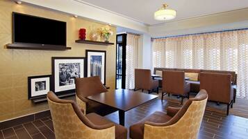 Un hall de hotel con zona de comedor, un televisor de pantalla plana montado en la pared, fotografías enmarcadas y sillas estampadas.