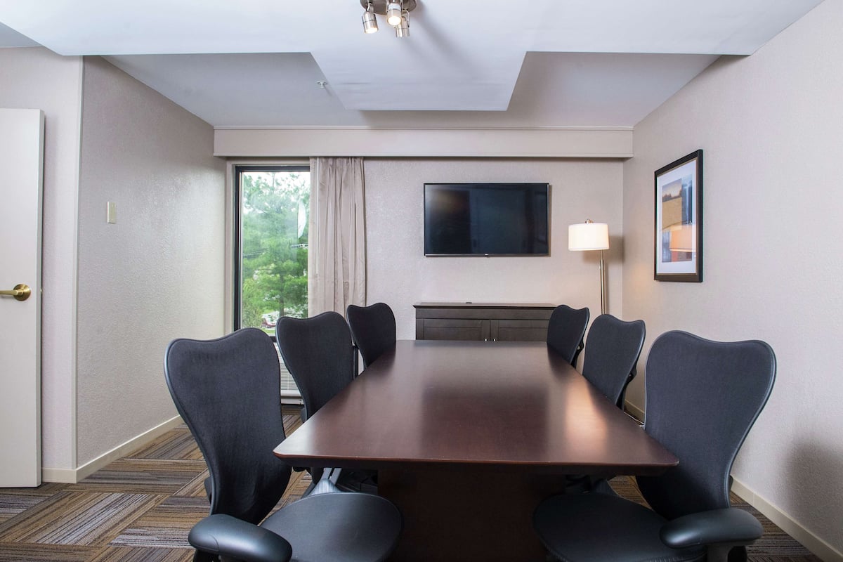 A conference room with a long table, six chairs, a flat-screen TV, a lamp, and a window with curtains.
