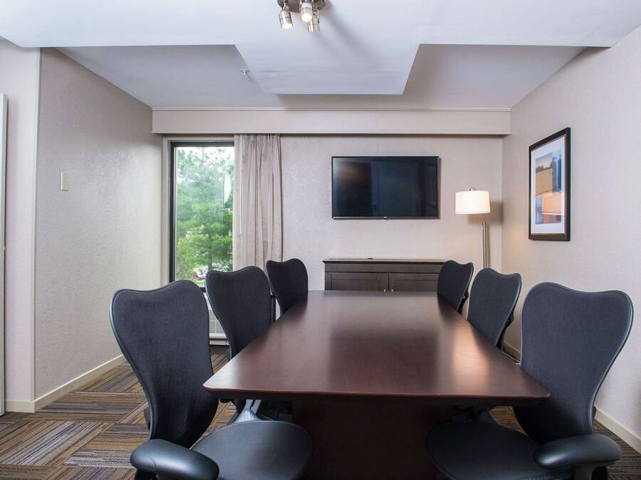 A conference room with a long table, six chairs, a flat-screen TV, a lamp, and a window with curtains.