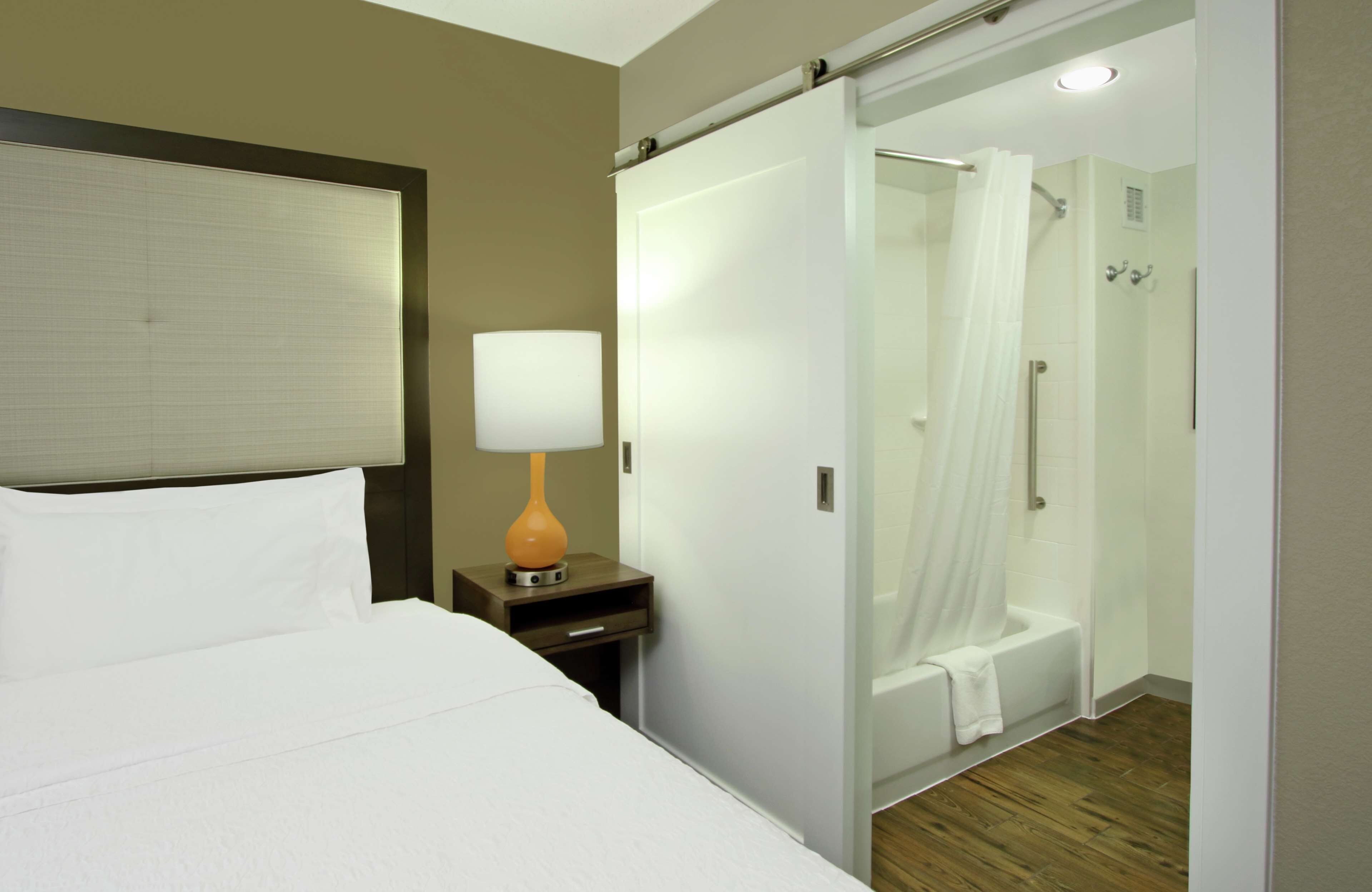 Una habitación de hotel con cama, una mesita de noche con lámpara, zona de ducha con puerta de cristal y suelo de madera.