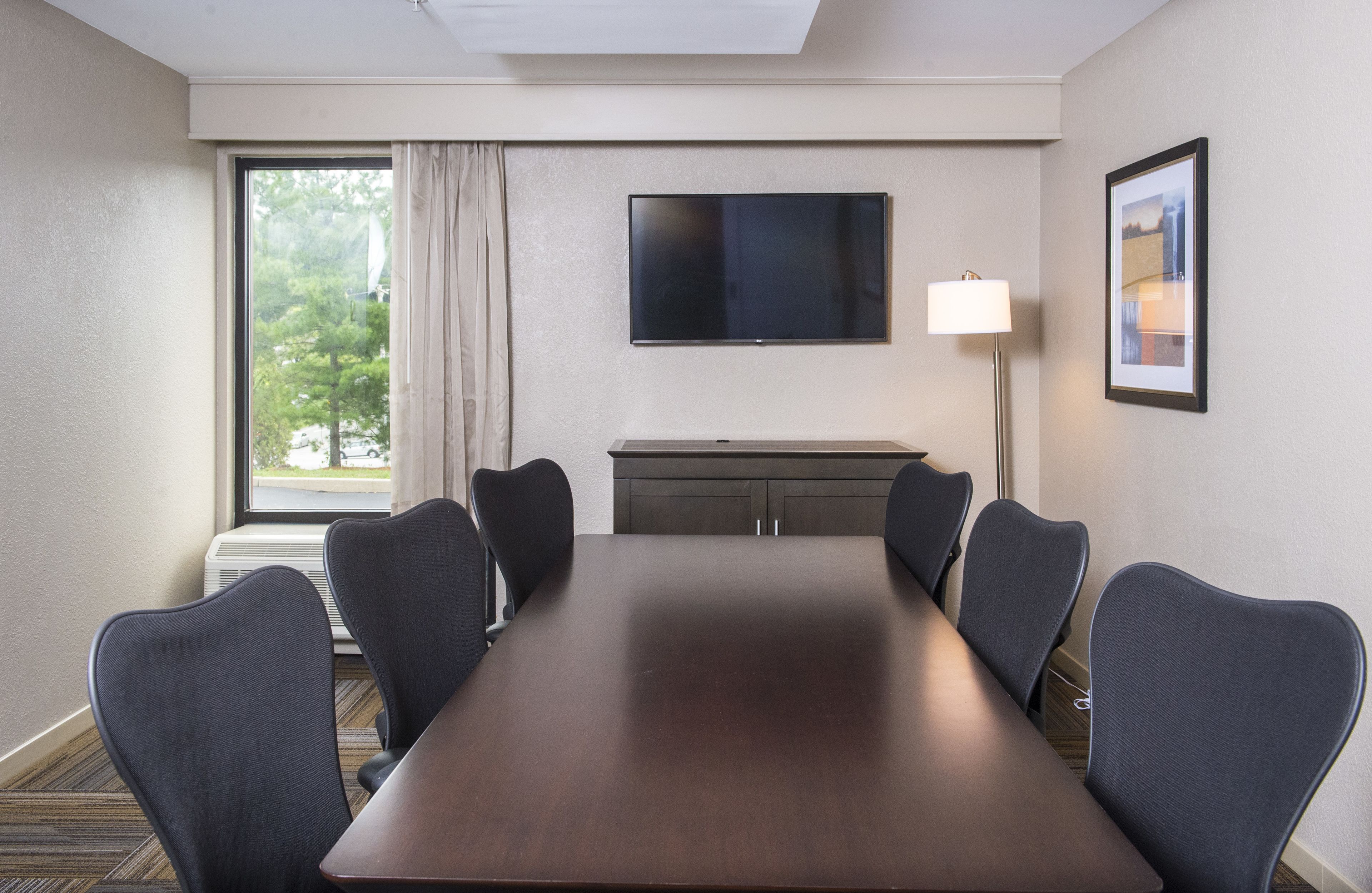 A conference room with a long table, six chairs, a flat-screen TV, a lamp, and a framed picture on the wall.