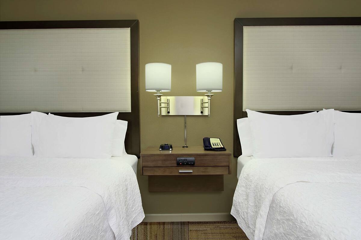 A hotel room with two beds, a nightstand with a lamp, a phone, and a small table.