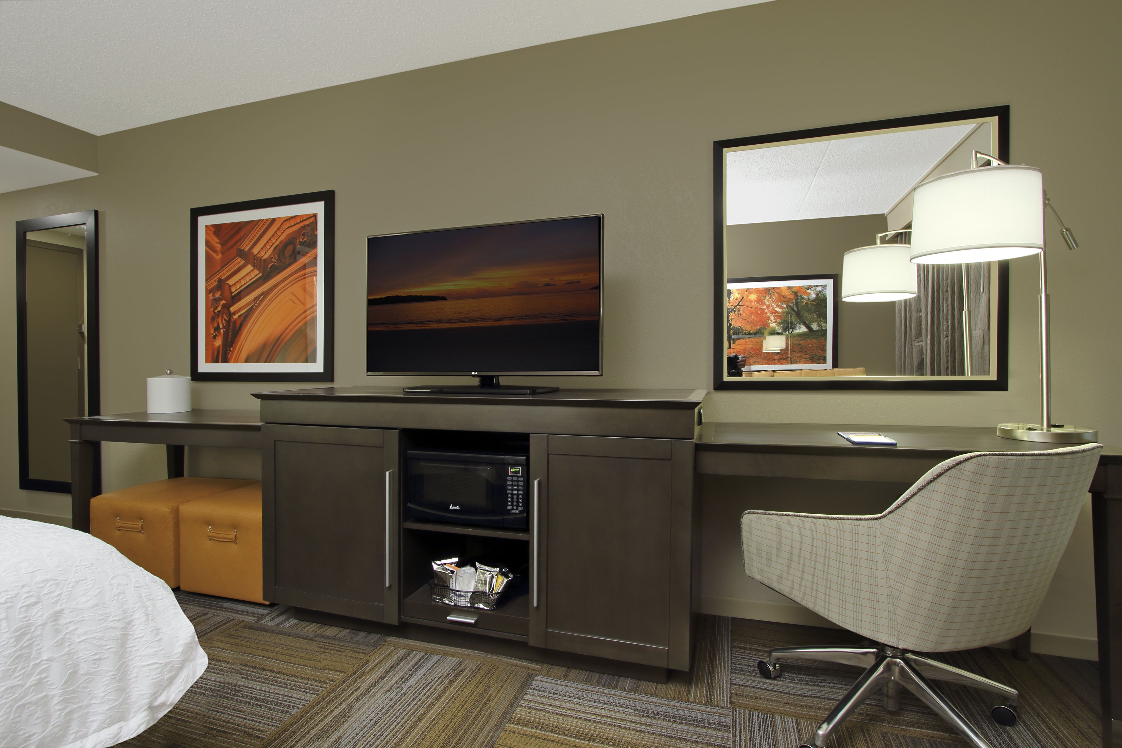 A hotel room with a bed, a desk with a flat-screen TV, a microwave, a chair, and a large mirror.