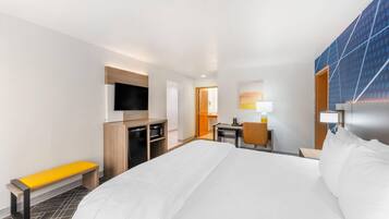 Quarto de hotel com uma cama grande, uma escrivaninha com cadeira, uma TV na parede, um banco e uma porta que dá para outro quarto.