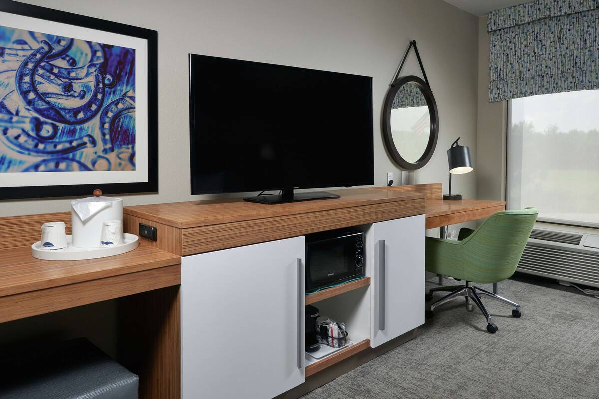 Habitación de hotel con escritorio de madera, televisor de pantalla plana, microondas, silla de oficina verde y una ventana grande.
