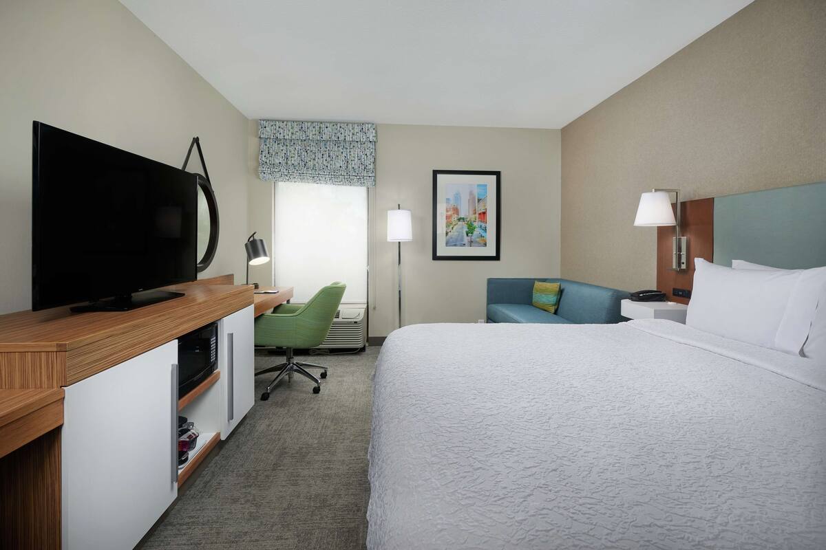 Una habitación de hotel con cama, escritorio con televisor de pantalla plana, una silla verde, sofá azul, un cuadro en la pared y ventana con cortina estampada.