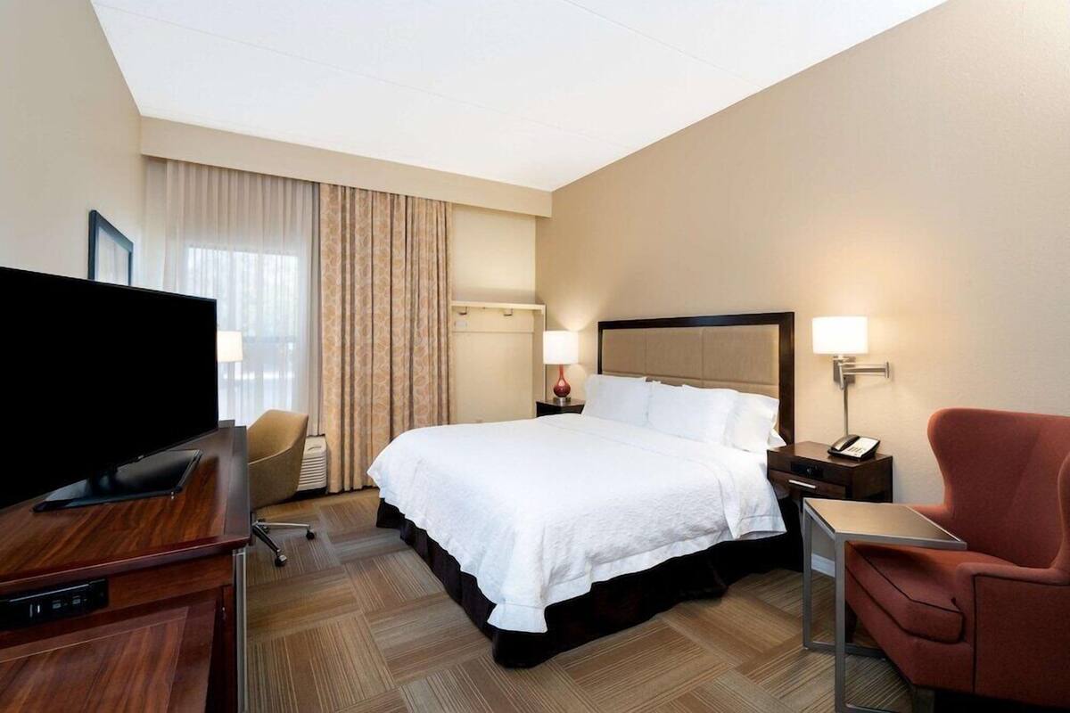 A hotel room with a large bed, a desk with a flat-screen TV, a chair, a bedside table with a lamp, and a window with curtains.