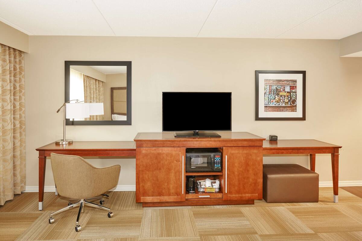 A hotel room with a wooden desk, a television, a cushioned chair, a microwave, and a framed picture on the wall.