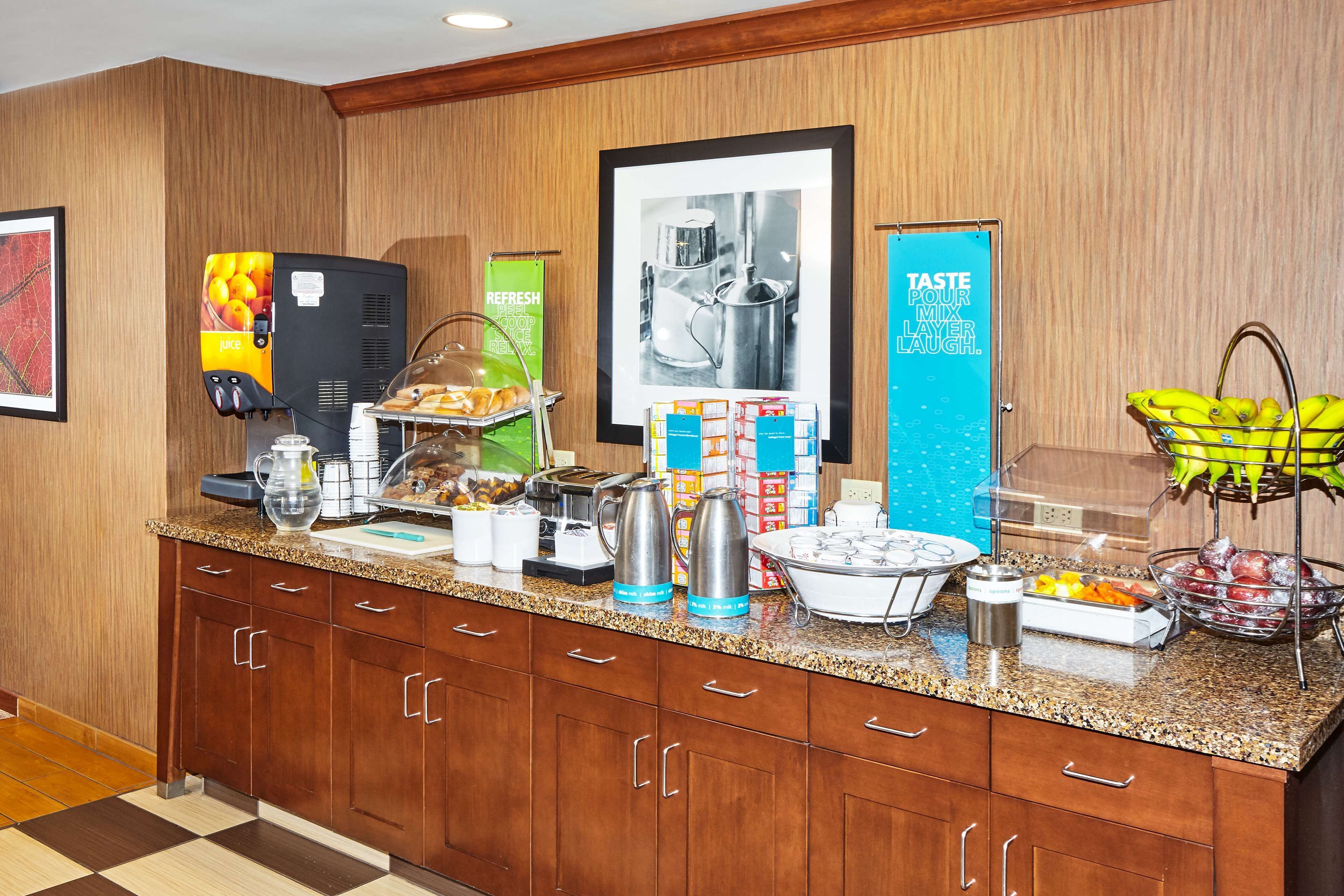 Un mostrador de desayuno de hotel con una máquina de café, dispensador de zumo y diversos alimentos.