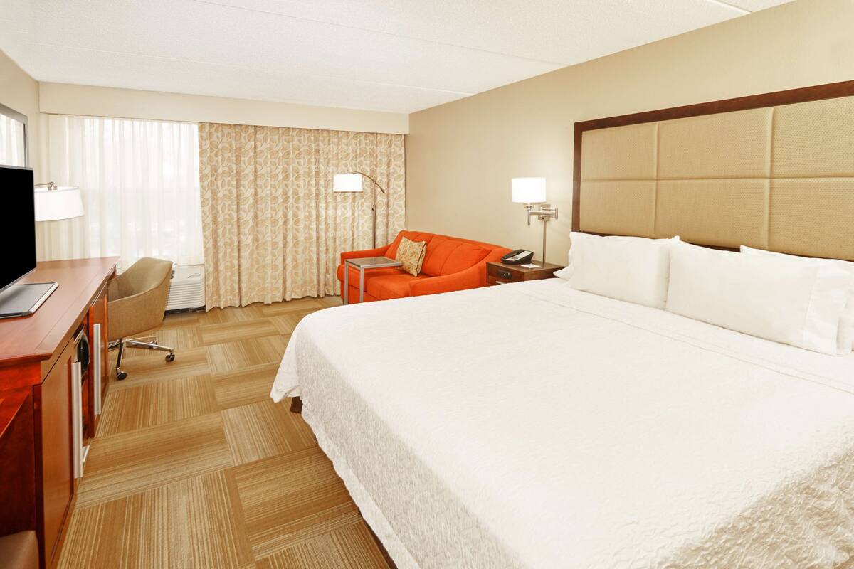 A hotel room with a large bed, a desk with a computer, an orange armchair, and a television.