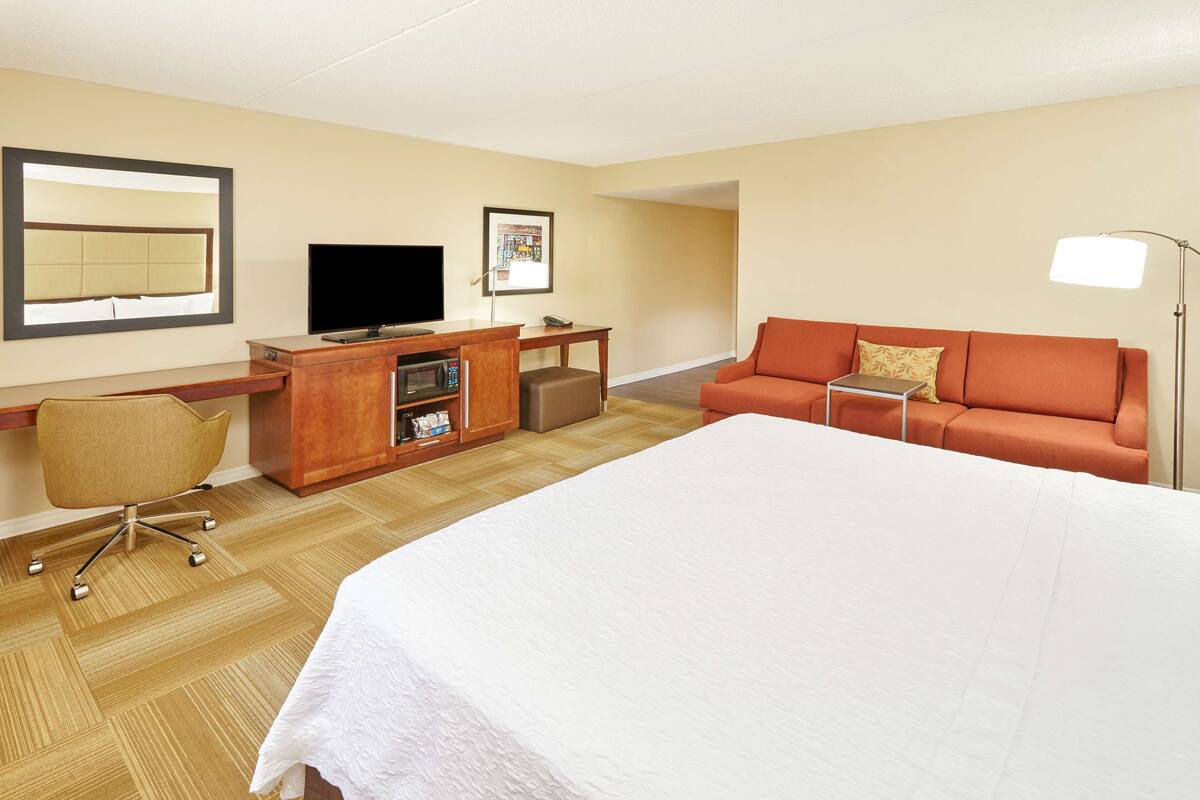 A hotel room with a large bed, a sofa, a desk with a television, a chair, and a lamp.