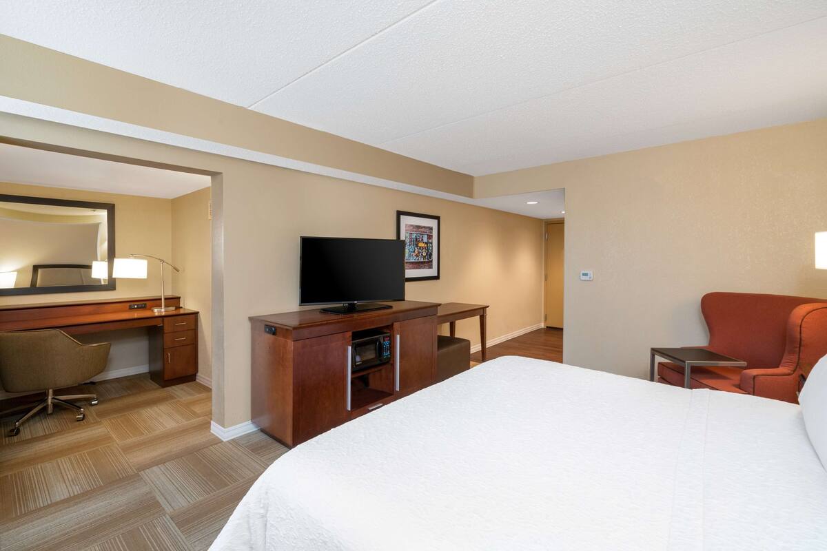 A hotel room with a large bed, a desk with a flat-screen TV, a chair, and a small table.
