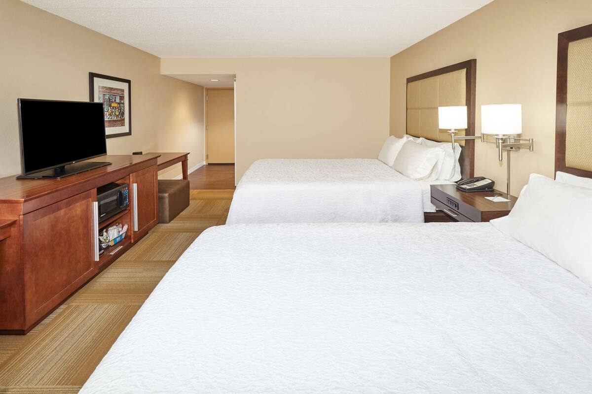 A hotel room with two beds, a television on a wooden cabinet, a desk, and a nightstand with a lamp.