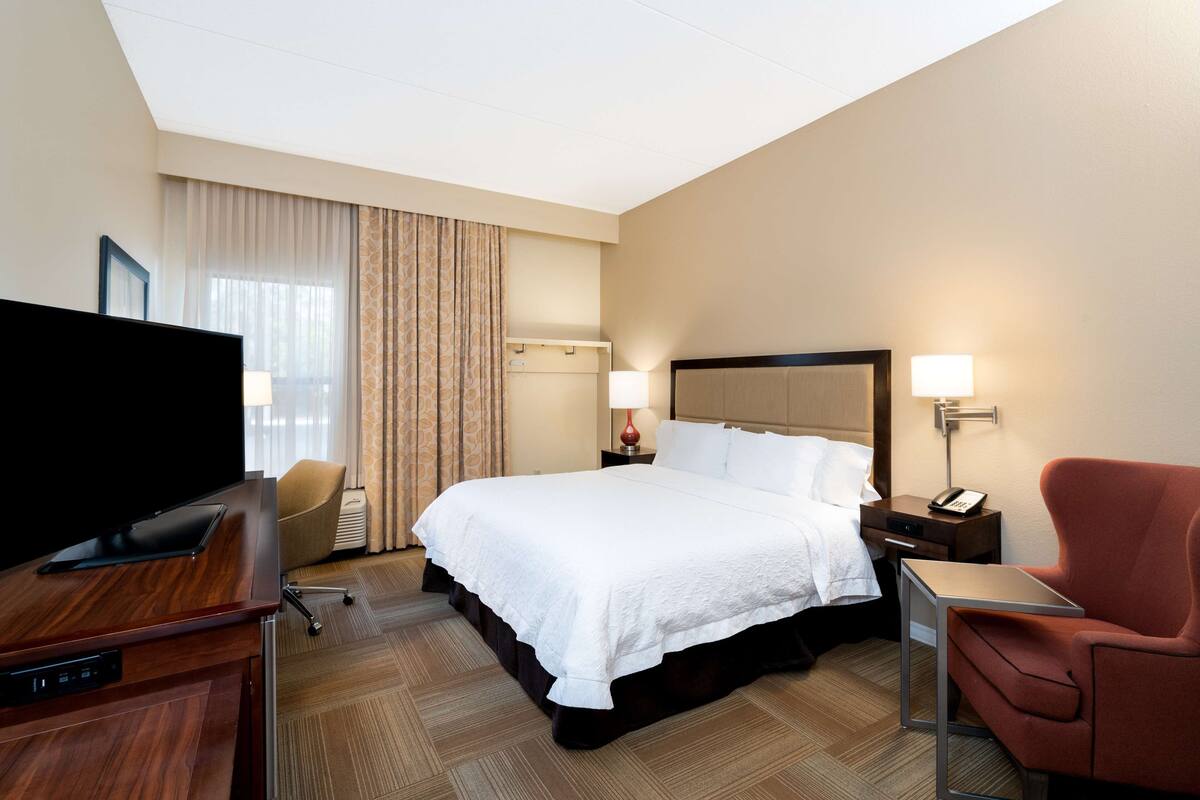 A hotel room with a large bed, a desk with a flat-screen TV, a chair, a bedside table with a lamp, and a window with curtains.