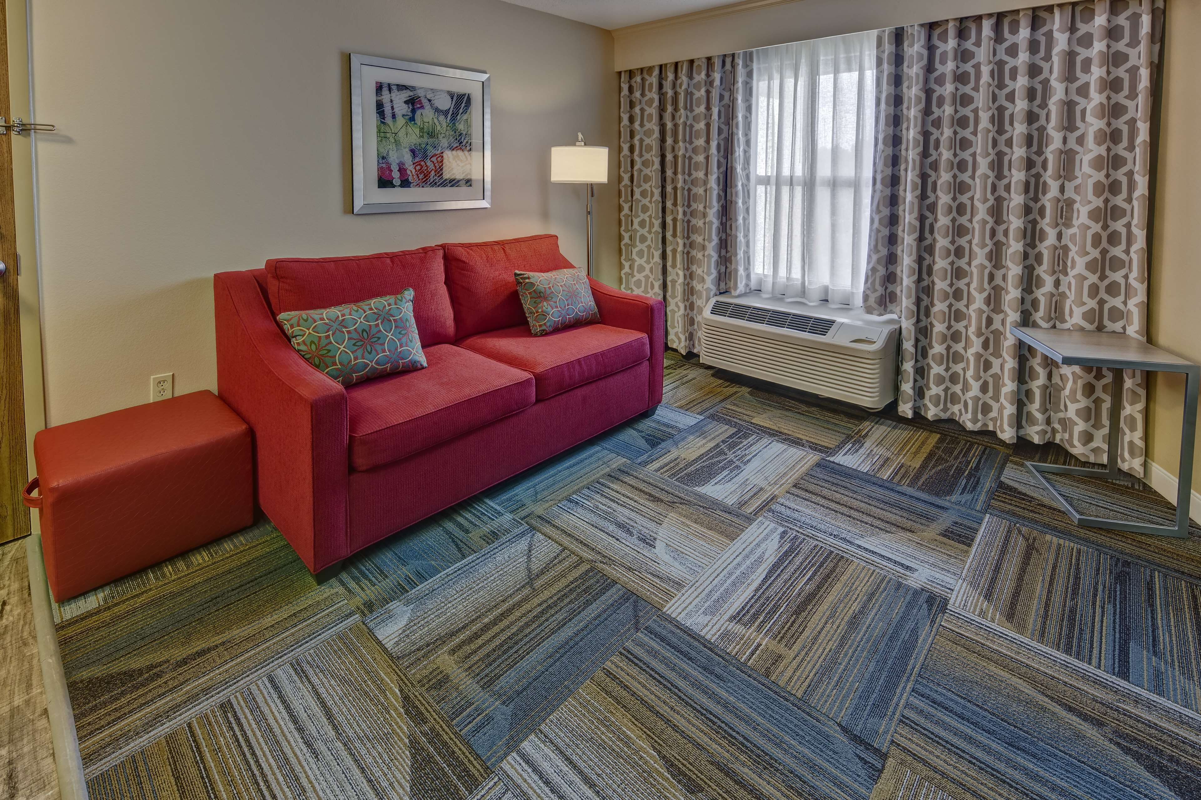 A hotel room with a red sofa, patterned carpet, a framed picture on the wall, a window with patterned curtains, and an air conditioning unit.