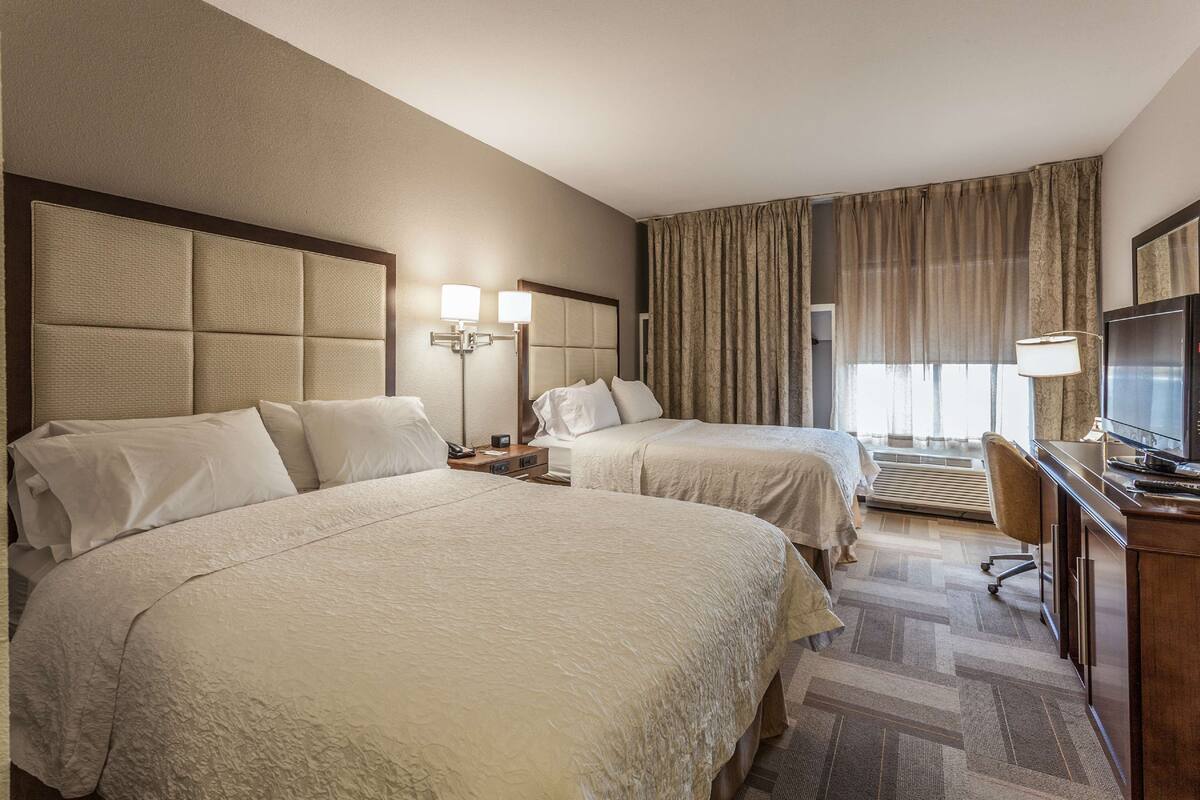 A hotel room with two beds, a desk with a television, and a large window with curtains.