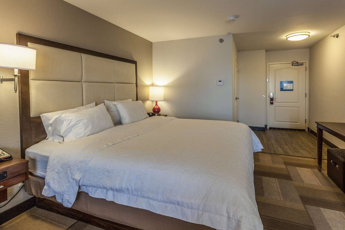 A hotel room with a large bed, a bedside table with a lamp, a desk, and a door.