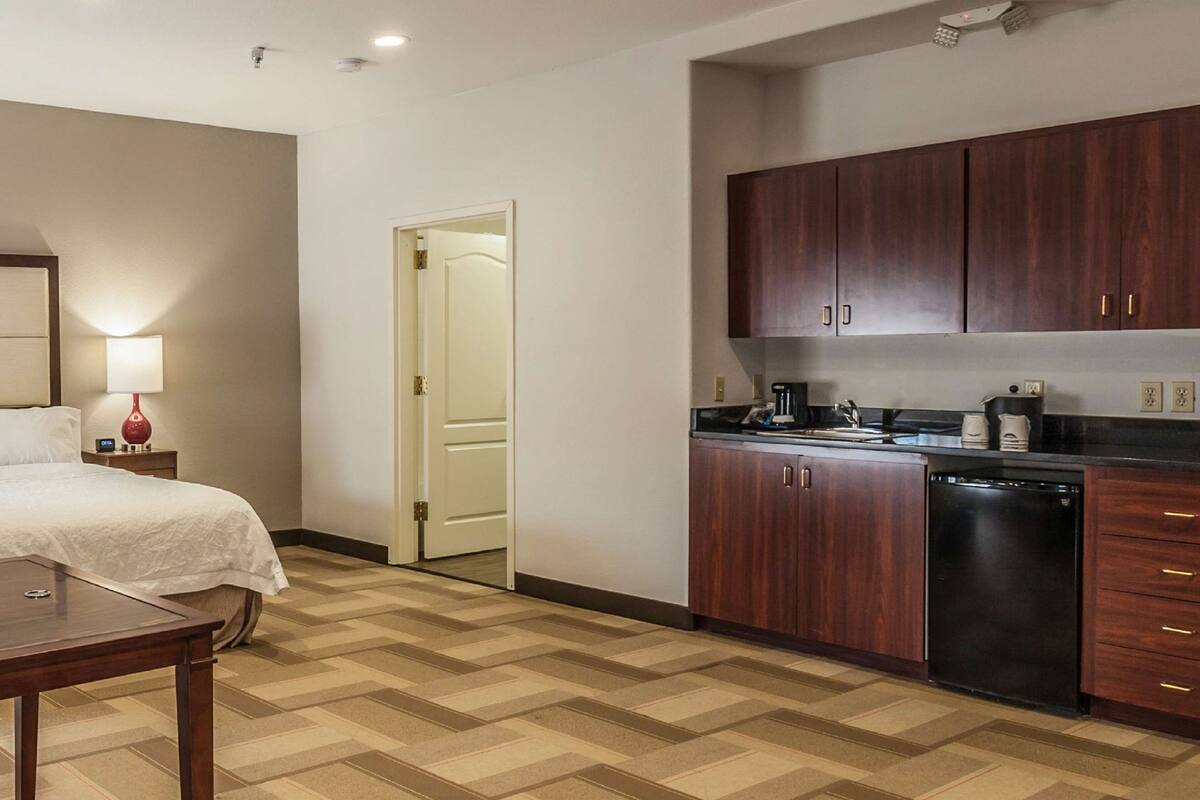 A hotel room with a bed, bedside table, lamp, and a kitchenette with cabinets and a dishwasher.