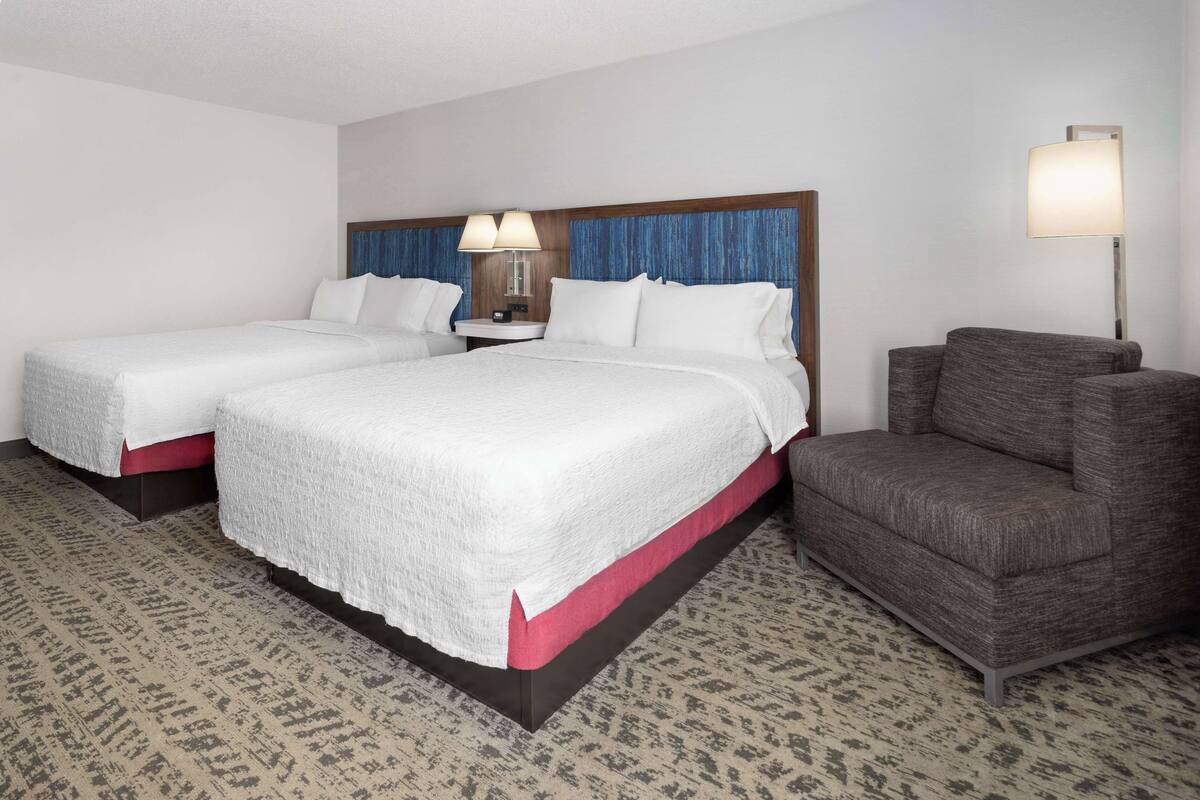 A hotel room with two beds, a nightstand, a lamp, and a sofa.