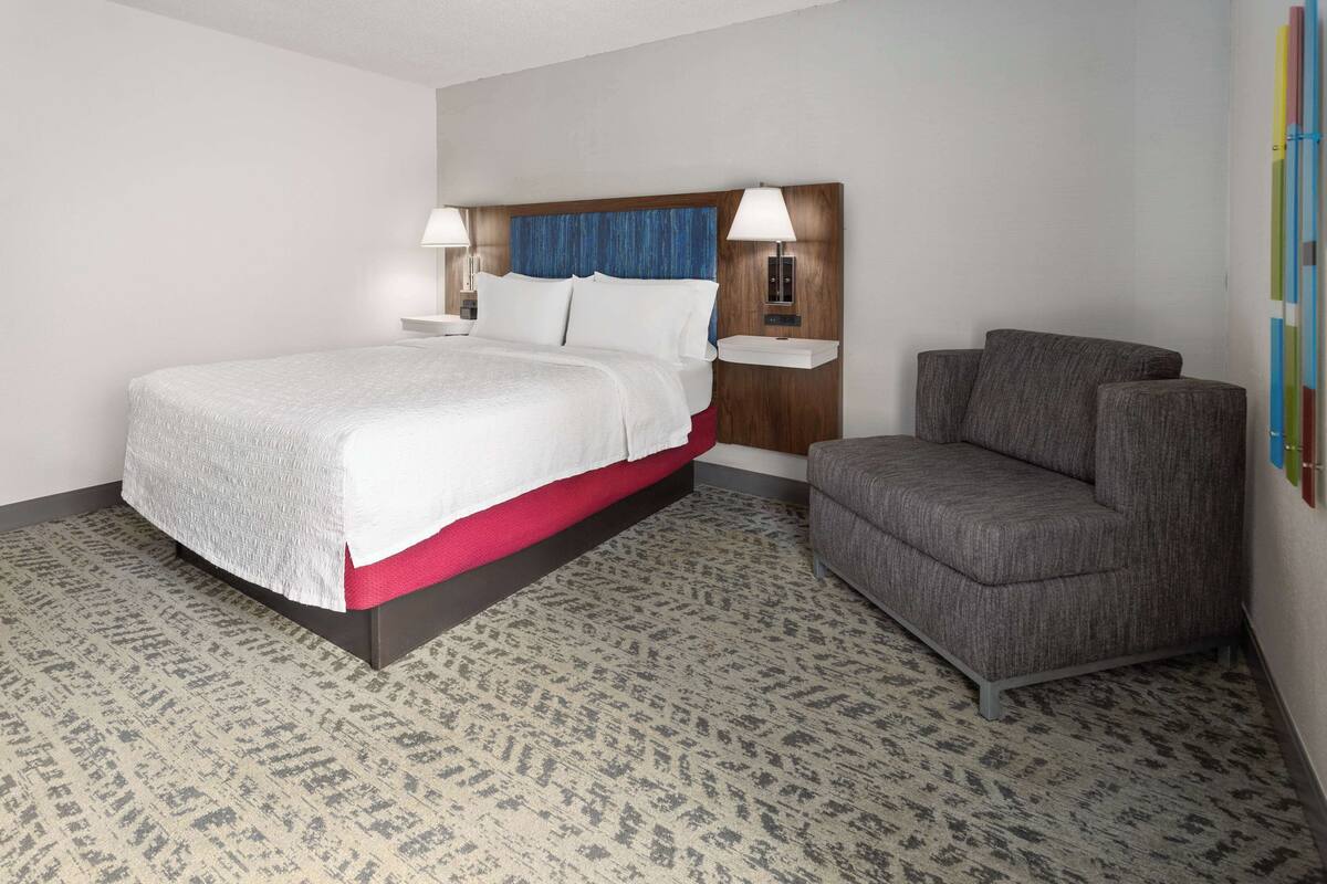 A hotel room with a large bed, a gray armchair, a nightstand with a lamp, and a wall-mounted shelf.