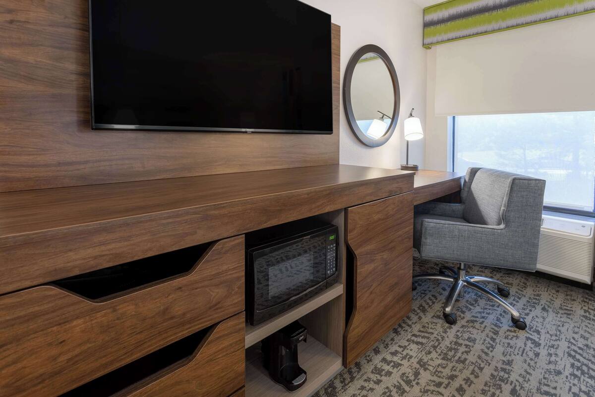 A modern hotel room with a flat-screen TV mounted on a wooden cabinet, a microwave, a coffee maker, a grey office chair, and a large window offering a view.